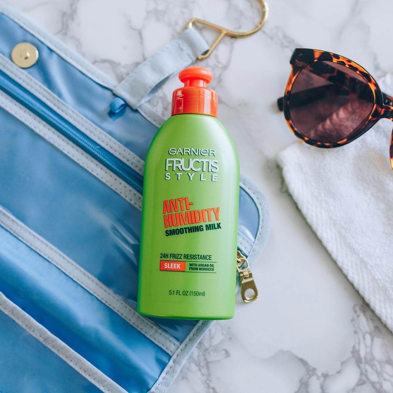 Garnier Fructis Style Anti-Humidity Smoothing Milk on a blue fold-out travel kit on white marble next to leopard-print sunglasses on a white wash cloth.
